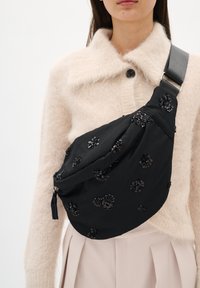 Fekete anyagú válltáska virágos flitter díszítésekkel, párnázott kialakítással és állítható pánttal. Keresztben viselve, világos pulóverrel párosítva.