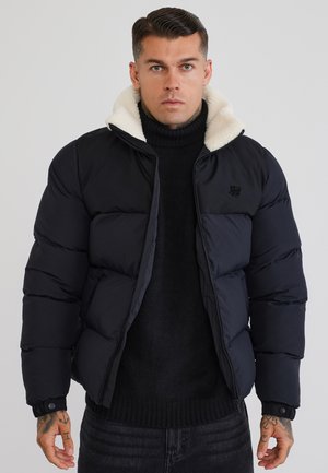 Uomo che indossa una giacca imbottita nera con colletto in pile bianco sopra un maglione dolcevita nero, in piedi davanti a uno sfondo grigio uniforme.