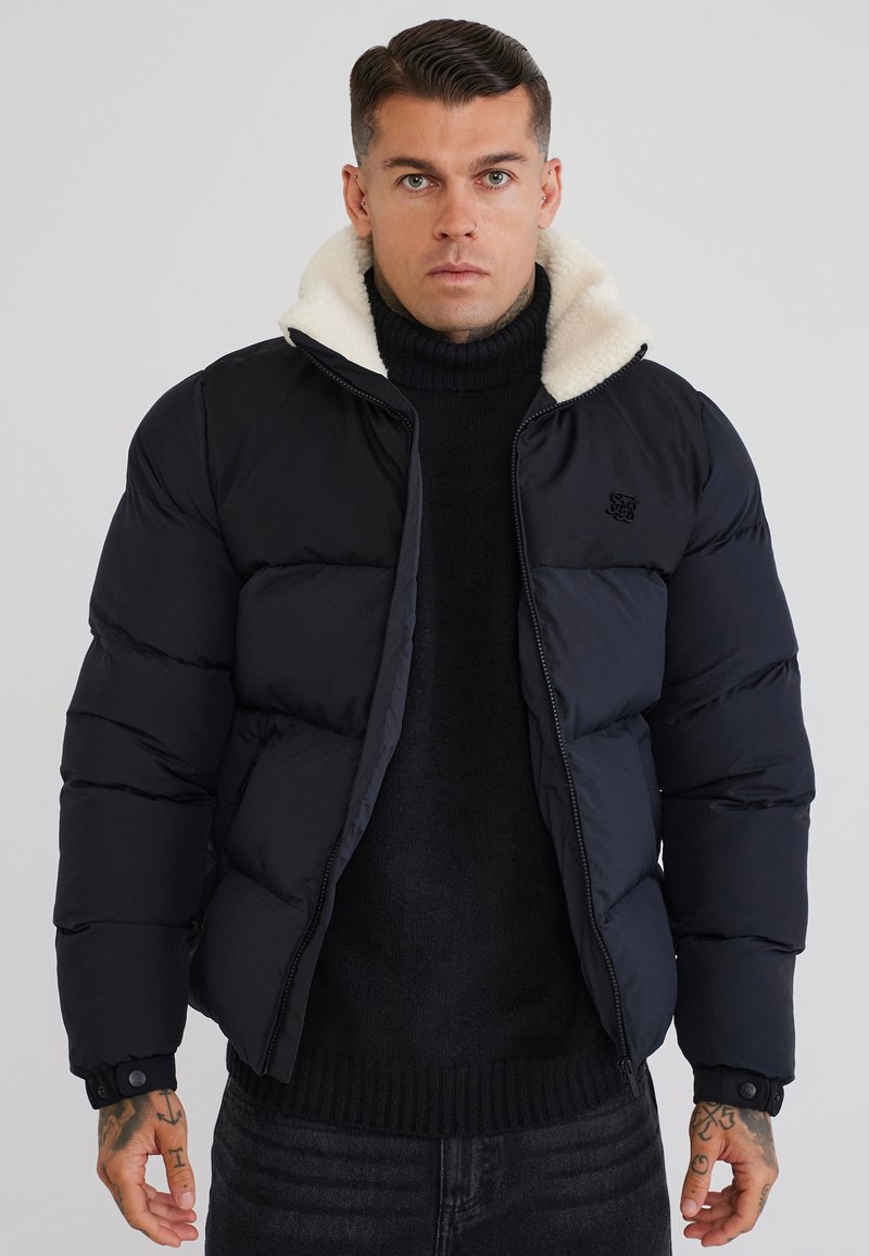 Άνδρας που φοράει μαύρο μπουφάν με λευκό fleece γιακά πάνω από μαύρο πουλόβερ ζιβάγκο, στέκεται μπροστά σε απλό γκρι φόντο.