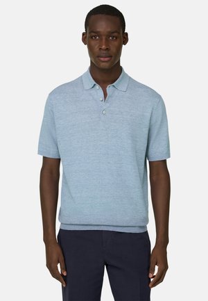 Mann trägt ein hellblaues Polohemd mit kurzen Ärmeln und eine dunkle Hose, steht vor einem schlichten hellen Hintergrund und blickt nach vorne.