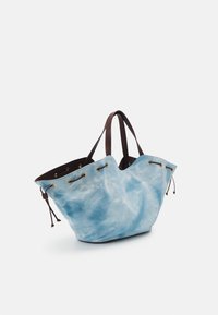 Sac fourre-tout en bleu clair tie-dye avec poignées en cuir marron foncé et œillets dorés, ouverture supérieure et détails à cordon sur les côtés.