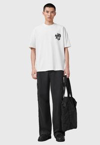 Camisa de algodón blanca con un logotipo gráfico negro en el pecho, combinada con pantalones cargo negros de pierna ancha y una bolsa tote negra.