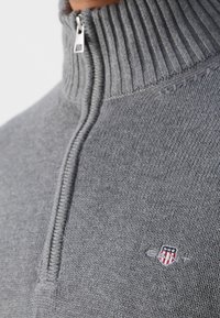 Gros plan d'un pull en maille côtelée gris avec un col zippé au quart et un petit logo brodé en forme de bouclier sur la poitrine.