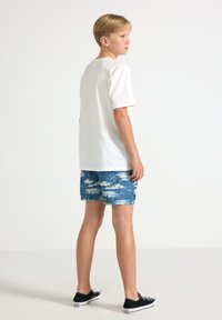 T-shirt bianco a maniche corte, pantaloni da bagno blu con motivo a palme, e sneaker nere in tela. Stile minimalista, vestibilità casual.