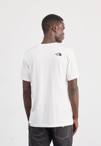 Bijela pamučna majica s okruglim izrezom i kratkim rukavima. Na gornjem dijelu leđa nalaze se crni logotip The North Face. Također su vidljive crne traperice.