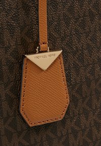 Brązowa skórzana torebka z metką w kształcie trójkąta z złotym metalowym akcentem z logo "Michael Kors", przymocowana brązowym paskiem. Tło przedstawia teksturowany materiał.