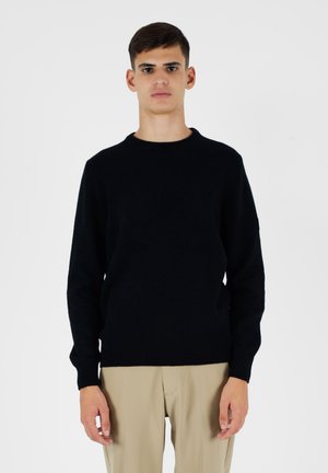 Maglione nero a maglia con scollo rotondo, maniche lunghe e polsini e orlo a costine. Tessuto liscio, design aderente, indossato sopra pantaloni beige.