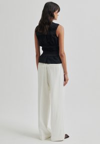 Juoda be rankovių maikutė su surinktu tekstūru ir kontrastingomis siūlėmis, derinama su laisvomis baltomis pločiais kelnais.