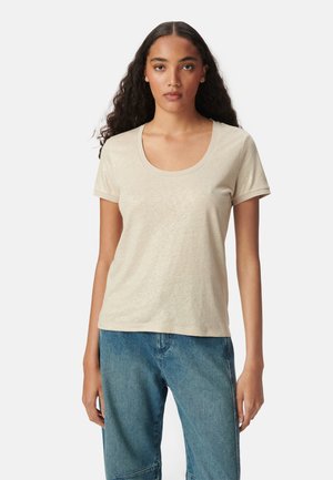 Giovane donna con capelli ricci che indossa una maglietta beige a maniche corte con scollo rotondo e jeans blu, in piedi contro uno sfondo bianco semplice.