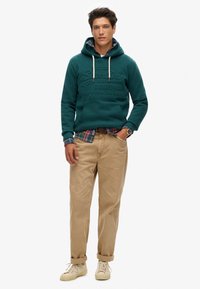Teal hoodie met getextureerde letters, beige chinos en lichte sneakers. Gelaagd met een geruite shirt zichtbaar bij de kraag en manchet.