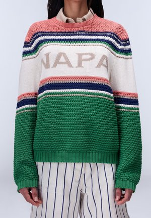 Person iført en farveblokstrikket sweater med teksten "NAPA", der har striber i pink, hvid, blå og grøn, kombineret med hvide bukser med tynde blå striber.