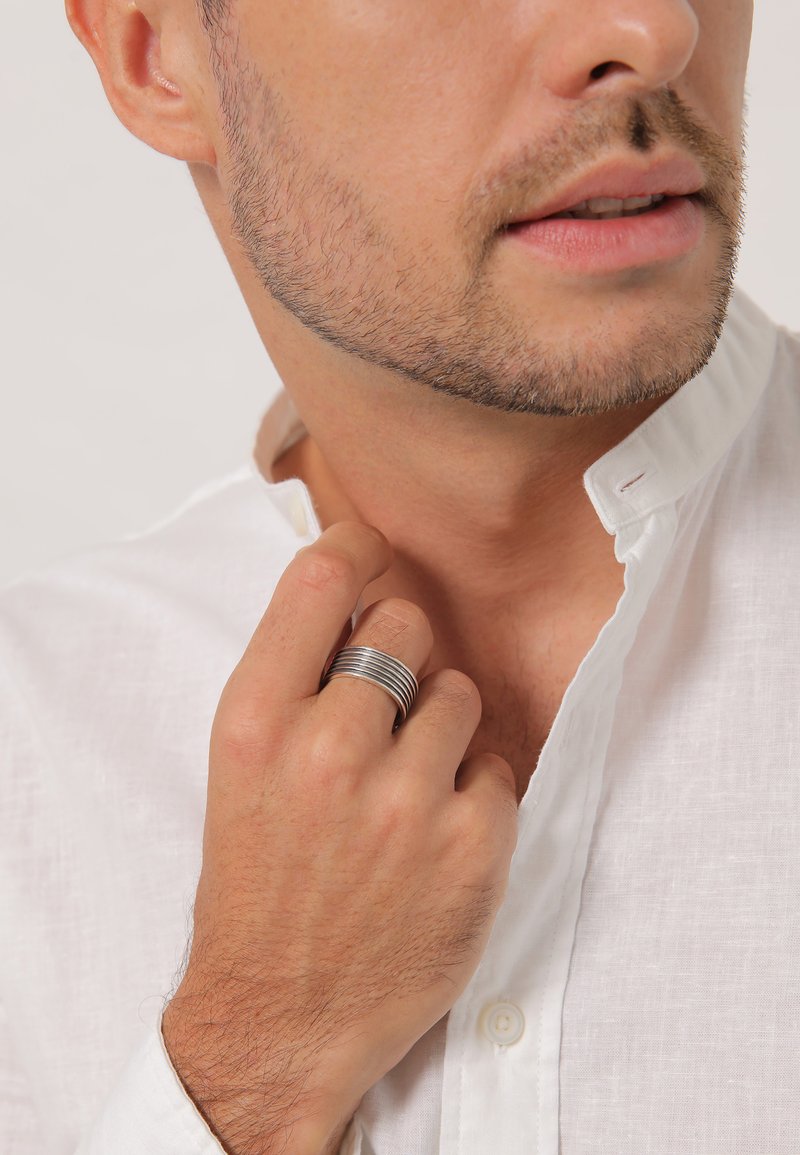 Metaling med stablede sølvbånd båret på en hånd nær en hvid skjortekrave. Nærbillede med fokus på hånden og ringens detaljer.