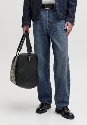 Άτομο που φοράει φαρδιά μπλε τζιν, μαύρα δερμάτινα παπούτσια, τζιν μπουφάν, κρατώντας μια μαύρη δερμάτινη τσάντα ταξιδίου από τις λαβές.