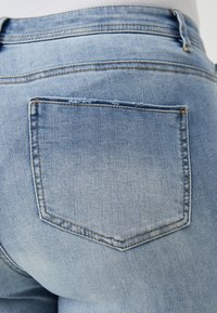 Světle modré džíny z denimu s obdélníkovou zadní kapsou, viditelným prošitím a hladkou texturou. Látka vykazuje jemné vyblednutí.