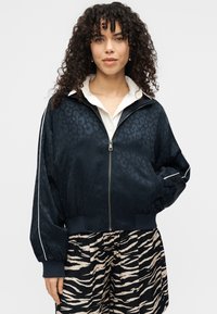 Μπουφάν bomber μπλε ναυτικό με λεοπάρ εκτύπωση, φερμουάρ μπροστά, ριμπ μανσέτες και γιακά, συνδυασμένο με μαύρο και μπεζ παντελόνι με ρίγες ζέβρας.