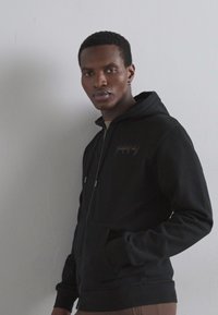 Hanorac negru cu fermoar, fabricat dintr-un material moale, cu buzunare frontale, glugă cu șnur și broderie subtilă a logo-ului pe piept. Croială structurată.