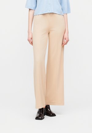 Kvinde iført beige bukser med brede ben, lyseblå cropped sweater med knapper og skinnende sorte ankelstøvler, stående foran en ensfarvet baggrund.