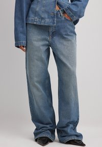 Šviesiai mėlyni laisvo kirpimo džinsai, pagaminti iš džinsinio audinio, su priekinėmis kišenėmis, mygtuko uždarymu ir šiek tiek apibrėžta senumo tekstūra. Stiliuoti su džinsiniu švarku.