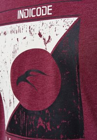 T-shirt in cotone color mogano con design grafico caratterizzato da un motivo triangolare in bianco e nero vissuto e un motivo circolare, etichettato "INDICODE."