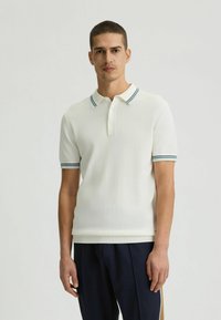 Weißes Poloshirt mit geripptem Kragen und blauen sowie grünen Streifen. Kurze Ärmel, schlanker Schnitt und weiche Textur. Getragen mit dunklen Hosen.