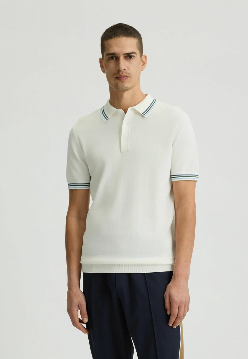 Weißes Poloshirt mit geripptem Kragen und blauen sowie grünen Streifen. Kurze Ärmel, schlanker Schnitt und weiche Textur. Getragen mit dunklen Hosen.