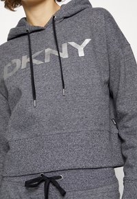 Šedá mikina DKNY z texturované látky, s velkým stříbrným logem, šňůrkami s kovovými konci a uvolněným střihem v pase.