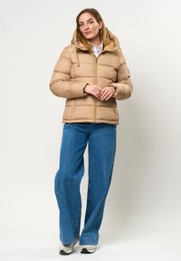 Béžová puffa bunda s kapucňou, so zapínaním vpredu a s jemným leskom. Skombinovaná s modrými džínsy s širokými nohavicami a športovými teniskami.