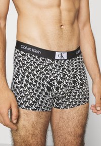 Calzoncillos boxer negros con un patrón de logotipos "CK" en blanco por toda la prenda y una cinturilla negra sólida con la marca "Calvin Klein".