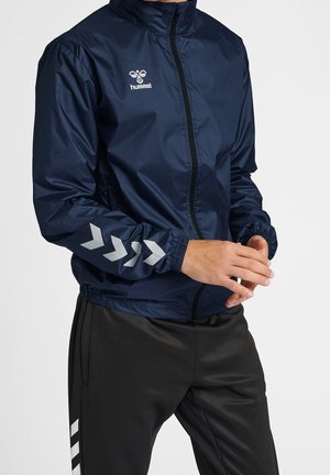 Veste de sport légère bleu marine avec fermeture éclair à l'avant, col montant, poignets élastiques et accents en chevron argentés sur les manches. Logo de la marque visible.