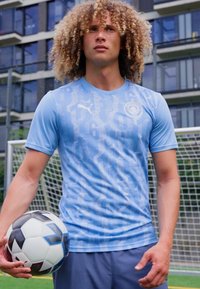 T-shirt de sport bleu clair à manches courtes, avec un motif numérique. Le joueur tient un ballon de football noir et blanc.