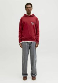 Rød hættejakke med en frontlomme, hvid tekstdesign, parret med grå lige bukser og brune sko. Simpelt, afslappet outfit.