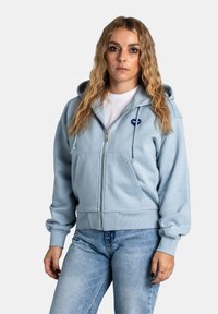 Felpa con zip di color azzurro chiaro, realizzata in tessuto morbido, con tasca frontale, cappuccio con cordino e logo ricamato sul petto.
