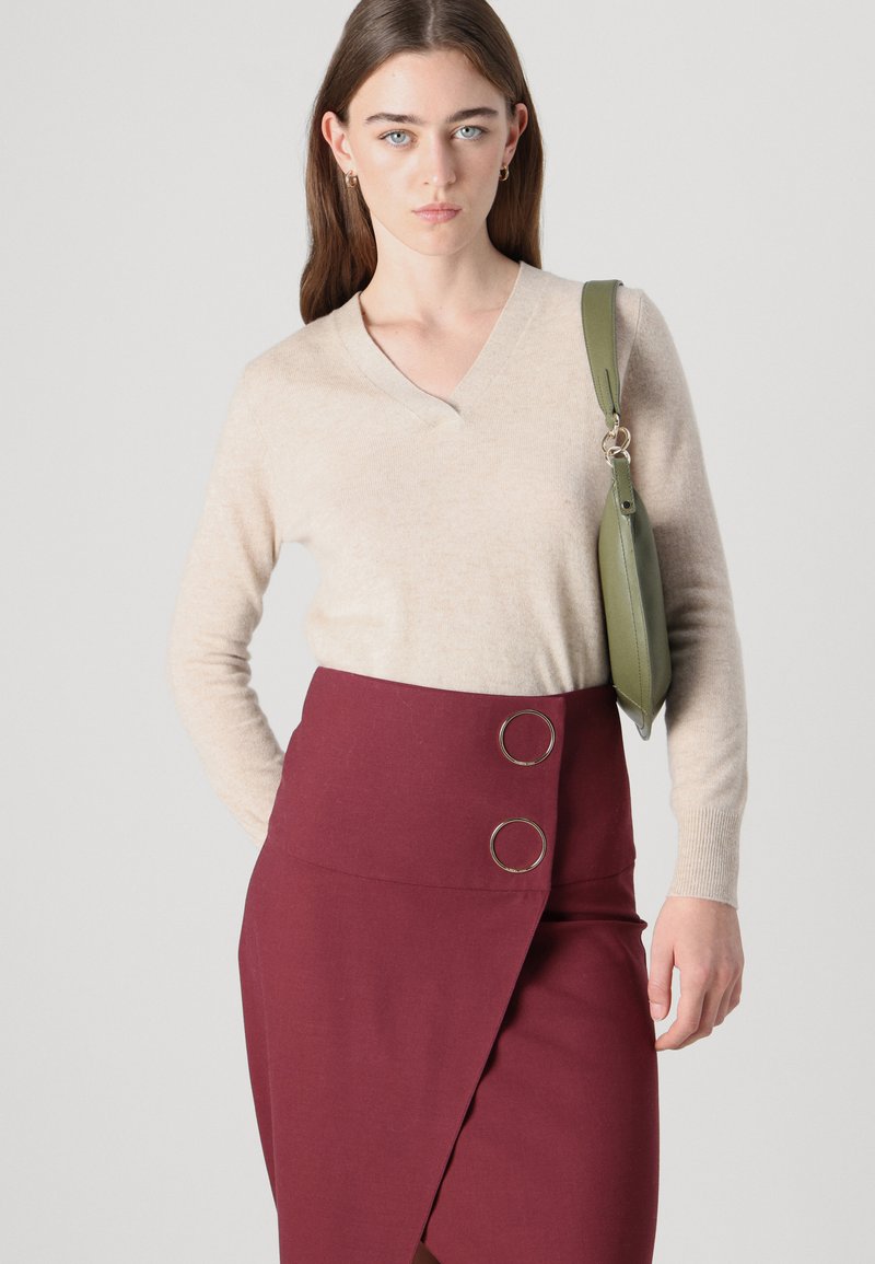 Beżowy sweter z kaszmiru w serek, zestawiony z bordową spódnicą kopertową z dwoma srebrnymi okrągłymi akcentami. Na ramieniu noszona jest zielona torebka.
