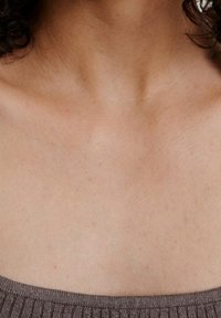 Närbild av en persons hals och övre bröst som bär en ribbad brun topp, med lockigt mörkt hår delvis synligt.