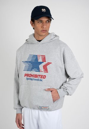 Mand iført grå hættetrøje med blå og rød stjerne-logo og teksten "PROHIBITED Sporting Goods Inc.", marineblå kasket med hvidt emblem, kigger til siden.