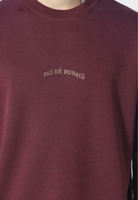 Burgundská bavlněná tričko s kulatým výstřihem. Na hrudi se nachází zlatě vyšitý nápis "PAS DÉ MONACO" ve zakřiveném uspořádání.