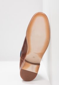 Chaussure en cuir marron avec design perforé, semelle beige et logo gravé. Taille 37 indiquée sous la chaussure. Forme de bout plat et arrondi.