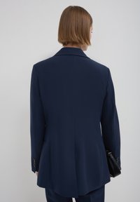 Blazer militare blu con colletto a punta, spalle strutturate e spacco posteriore singolo. Presenta polsini con bottone e una texture di tessuto liscia.