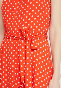 Röd tyg med vita prickar, med en knut i midjan. Slät textur, ärmlös design och A-linjeformad siluett.