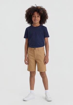 Garçon aux cheveux bouclés portant un t-shirt bleu marine, un short beige jusqu'aux genoux, des chaussettes blanches et des baskets blanches, se tenant devant un fond uni.