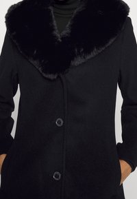 Černý vlněný kabát s velkým límcem z umělé kožešiny, dvěma knoflíky a bočními kapsami; hladká textura s čistým, strukturovaným designem.