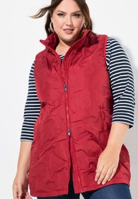 Rote gesteppte Weste mit hohem Kragen, Reißverschluss vorne und zwei Seitentaschen. Über einem schwarz-weiß gestreiften Langarmshirt getragen.