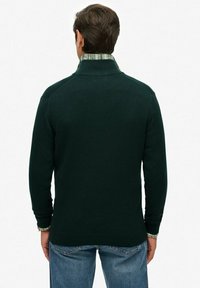 Suéter verde oscuro con textura acanalada y un cuello alto, superpuesto a una camisa de cuadros. Costuras visibles y ajuste ceñido en los puños.