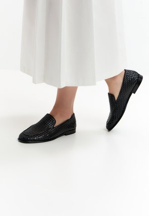 Schwarze gewebte Slip-On Schuhe, getragen zu einem weißen Rock, zeigen Füße, wobei ein Fuß leicht auf einem weißen Hintergrund angehoben ist.