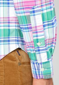 Flerfärgad rutig skjorta med grönt, rosa, blått och vitt samt knappmanschetter; kombinerad med ljusbruna manchesterbyxor.