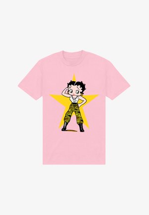 Personnage aux cheveux noirs courts portant un haut blanc, un pantalon camouflage, des bottes noires, des boucles d'oreilles dorées, posant avec une main sur la tête devant une étoile jaune sur un t-shirt rose.