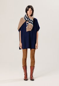 Navyblauwe poncho-trui, beige en navy gestreepte sjaal eroverheen gedrapeerd. Bruine suède leggings en kniehoge bruine leren laarzen. Eenvoudig ontwerp, relaxte pasvorm.