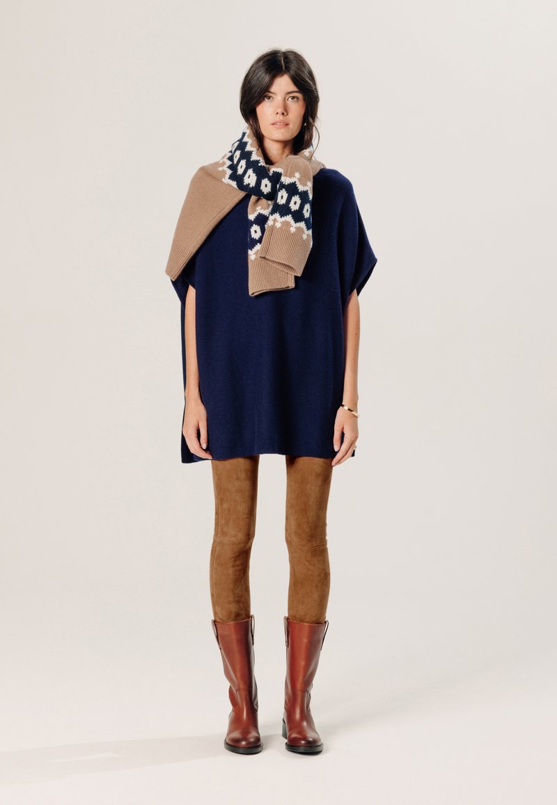 Navyblauwe poncho-trui, beige en navy gestreepte sjaal eroverheen gedrapeerd. Bruine suède leggings en kniehoge bruine leren laarzen. Eenvoudig ontwerp, relaxte pasvorm.