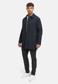 Navyblau wasserfeste Jacke mit einem Frontreißverschluss, zwei Seitentaschen und einem Kragen, kombiniert mit dunklen Hosen und weißen Sneakers.