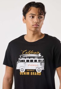 Svart T-shirt med en vintage Volkswagen-buss i vitt och orange, med texten "California dreaming" och "HIGH QUALITY DENIM BRAND".